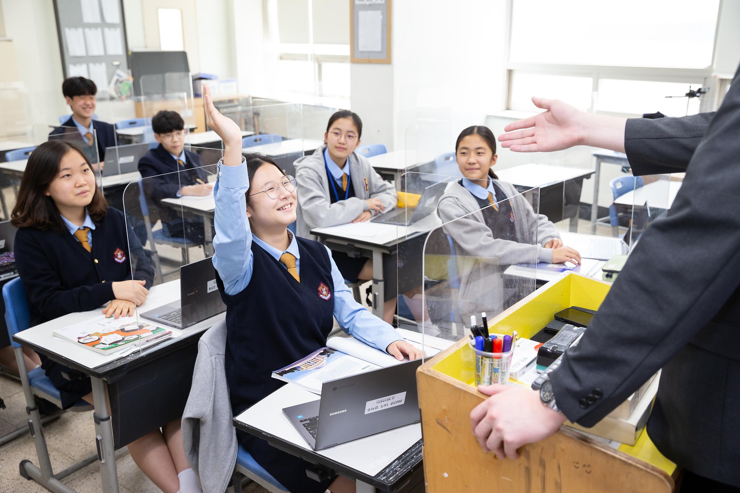 책상을 물리고 바닥에 앉아 영어 수업 중인 13명의 학생들. 교복을 입고 선생님의 동작을 따라하고 있다.